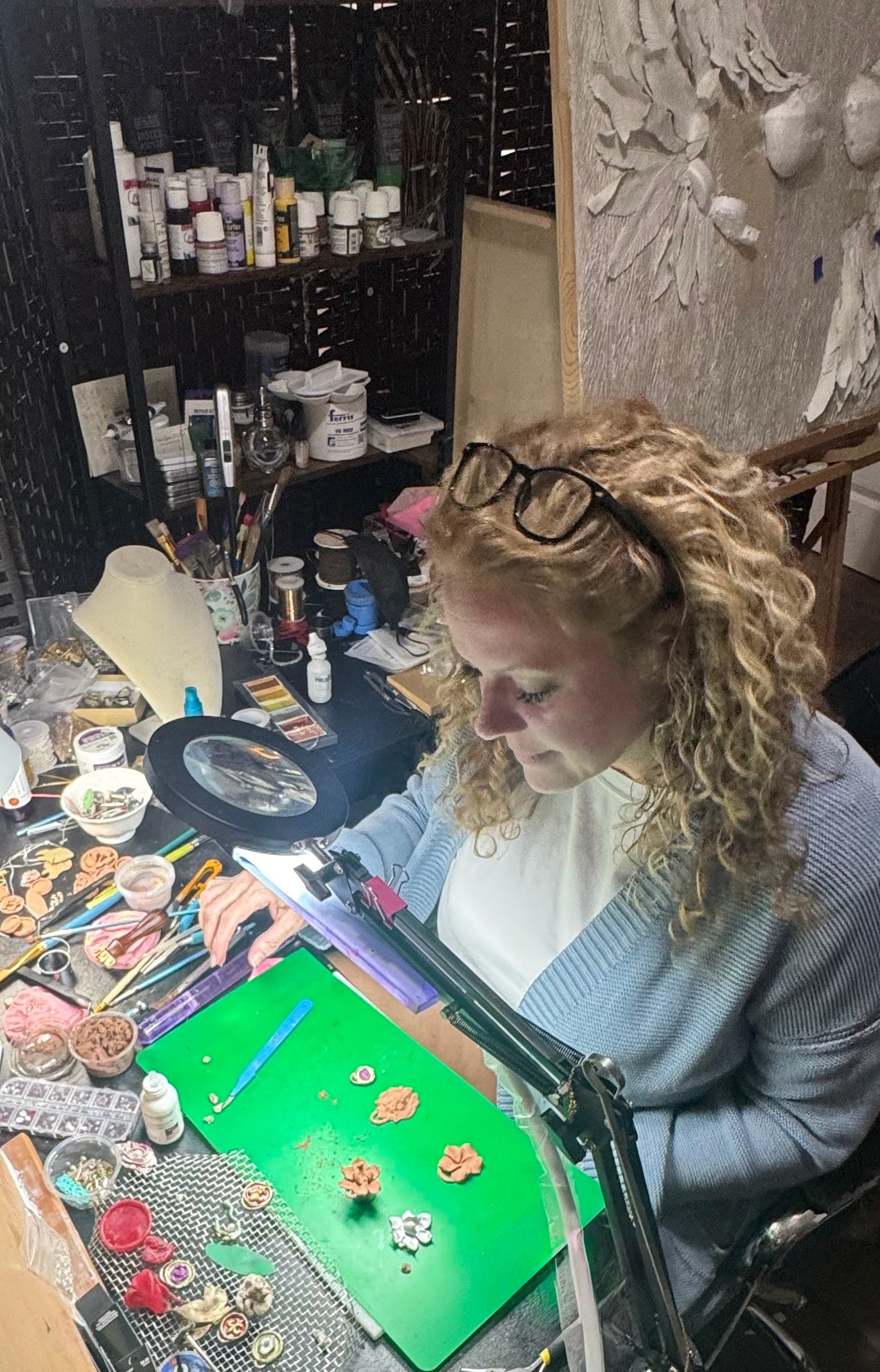 A woman with curly blonde hair and glasses sits at a cluttered craft desk, working under a bright lamp. The desk is covered in various small craft supplies, beads, and tools, and the background shows shelves filled with paintbrushes and art materials.